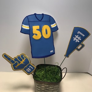 May include: A blue and gold football jersey with the number 50, a blue and gold foam finger with the number 1, and a blue and gold megaphone with the number 1, all in a metal bucket with green moss.