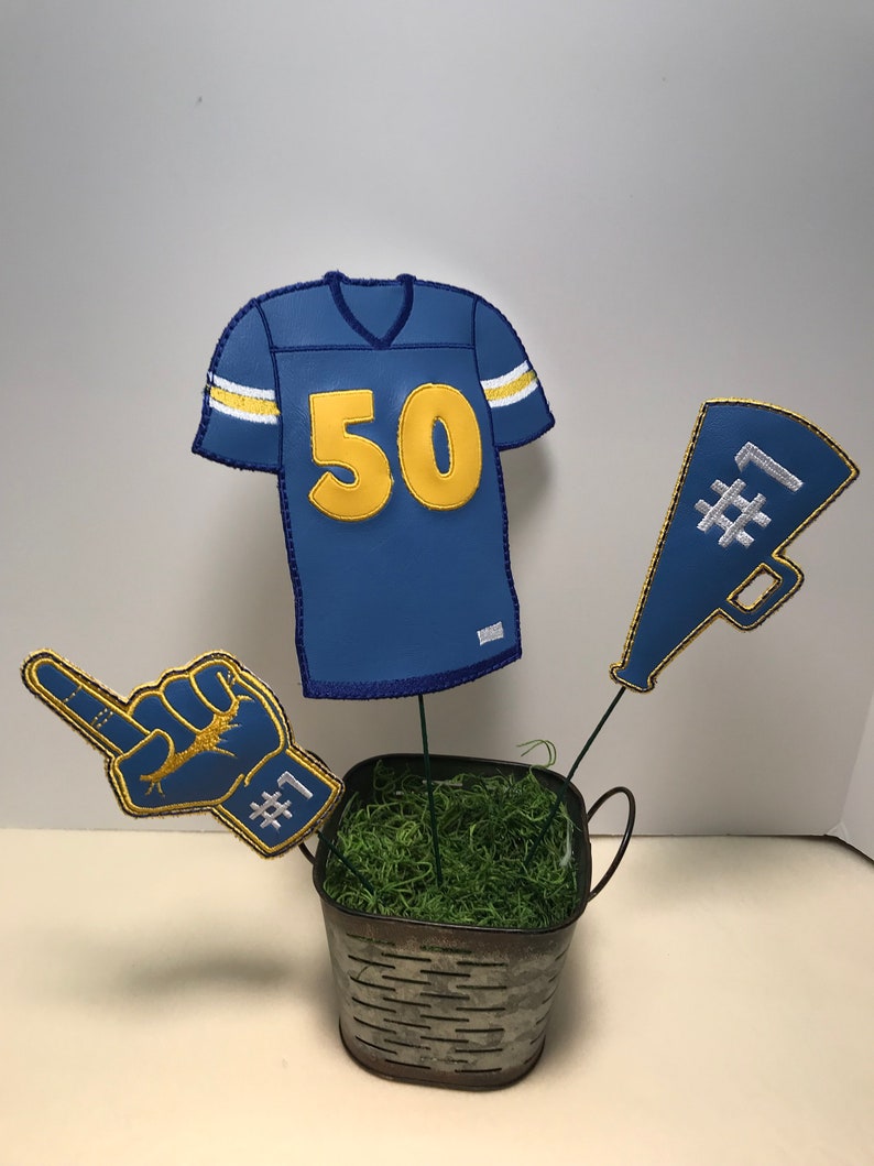 May include: A blue and gold football jersey with the number 50, a blue and gold foam finger with the number 1, and a blue and gold megaphone with the number 1, all in a metal bucket with green moss.