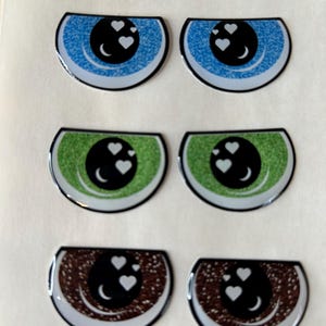 以下が含まれることがあります： 青、緑、茶色の半円形をした6つの目のデザイン。各目は、白いハートのアクセントと白い三日月が付いた黒い瞳を持っています。目は光沢のある仕上がりで、白い縁取りがあります。