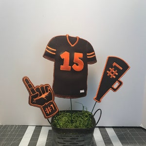 May include: A brown and orange football jersey with the number 15 on the front, a brown and orange foam finger with the number 1 on it, and a brown and orange megaphone with the number 1 on it. All three items are arranged in a metal bucket with green moss.