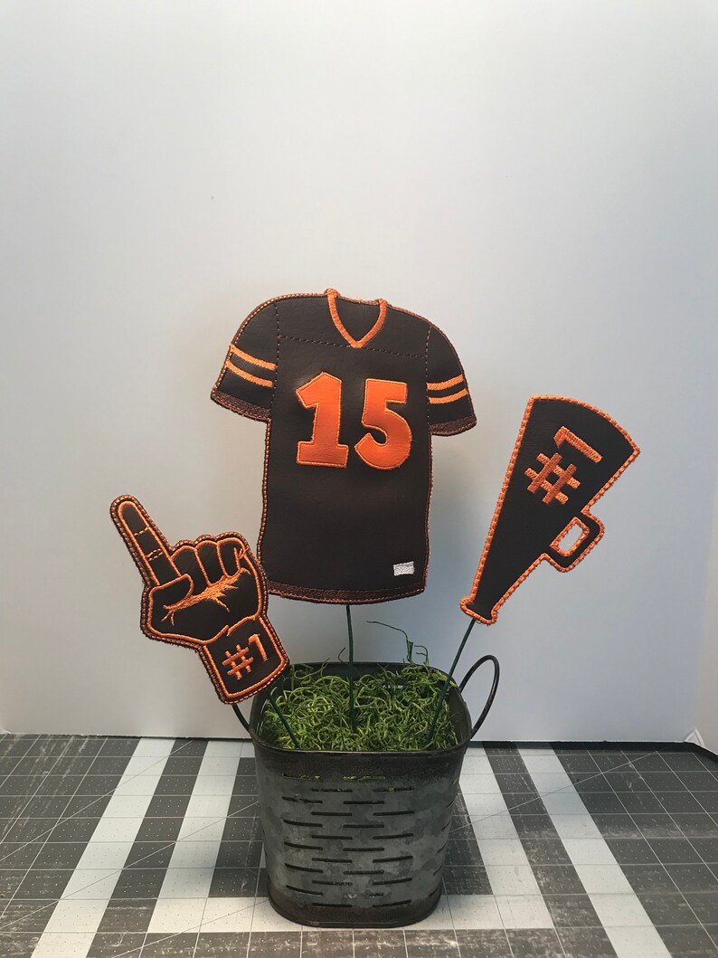 May include: A brown and orange football jersey with the number 15 on the front, a brown and orange foam finger with the number 1 on it, and a brown and orange megaphone with the number 1 on it. All three items are arranged in a metal bucket with green moss.