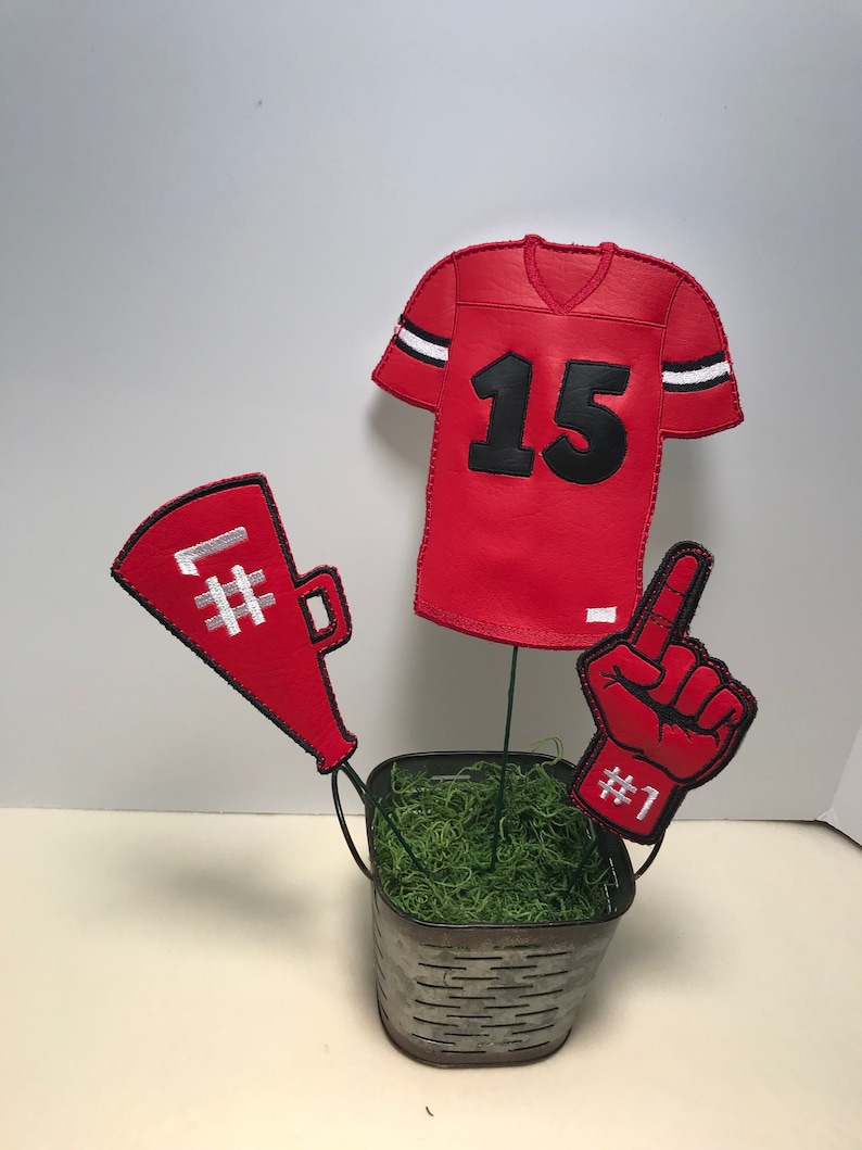 May include: A red and white football themed centerpiece with a jersey, a foam finger, and a megaphone. The centerpiece is in a small metal bucket with green moss.
