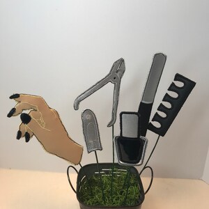 K&ouml;nnte beinhalten: Ein Eimer aus Metall, gef&uuml;llt mit gr&uuml;nem Moos und oben mit einer Hand und verschiedenen Nagelpflegewerkzeugen, darunter ein Nagelknipser, eine Nagelfeile, eine Flasche Nagellack und ein Kamm.