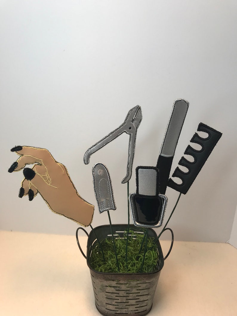 K&ouml;nnte beinhalten: Ein Eimer aus Metall, gef&uuml;llt mit gr&uuml;nem Moos und oben mit einer Hand und verschiedenen Nagelpflegewerkzeugen, darunter ein Nagelknipser, eine Nagelfeile, eine Flasche Nagellack und ein Kamm.