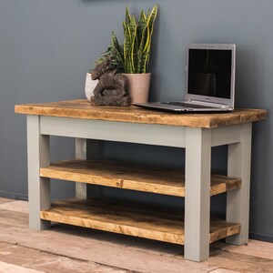 May include: A rustic wooden shelf unit with two shelves and a light gray painted finish. The shelf unit is made of reclaimed wood and has a natural wood grain. It is shown with a laptop computer and a potted plant on top.