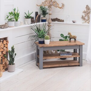 May include: A gray wooden shelf unit with two shelves. The shelf unit is made of reclaimed wood and has a rustic look. The shelf unit is sitting on a white floor in a room with plants and other decor.