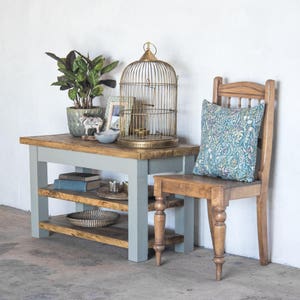 May include: A light gray wooden table with two shelves and a birdcage on top. The table is next to a wooden chair with a floral patterned cushion.