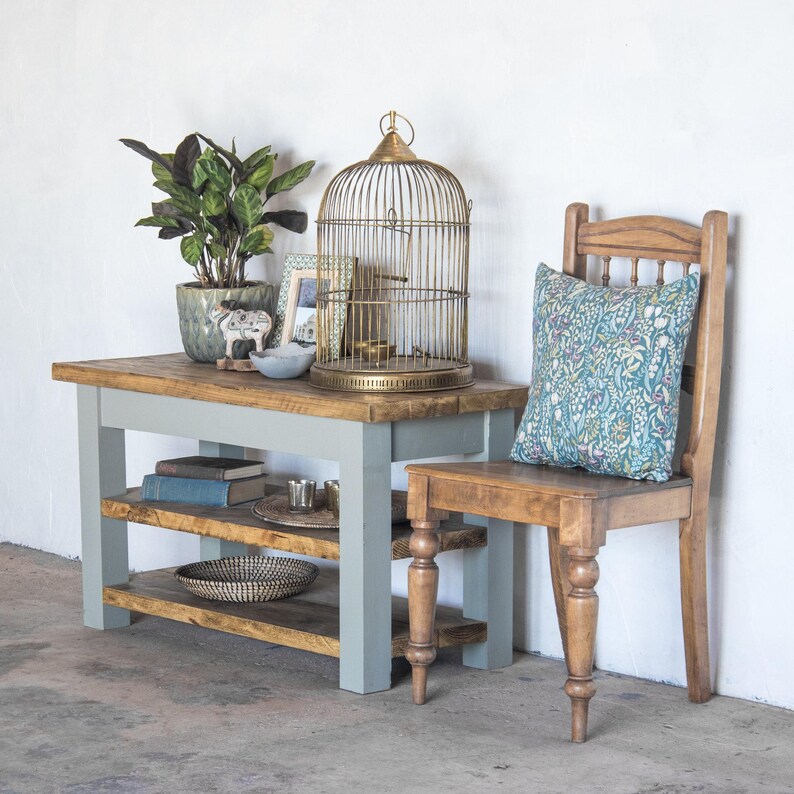 May include: A light gray wooden table with two shelves and a birdcage on top. The table is next to a wooden chair with a floral patterned cushion.