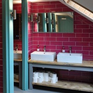 May include: Two white porcelain sinks with chrome faucets are mounted on a wooden countertop with a gray base. The sinks are in a bathroom with pink tile walls and a mirror above the sinks.