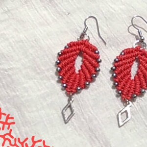 Pode incluir: Um par de brincos vermelhos e prateados com um pingente em forma de diamante. Os brincos s&atilde;o feitos com um cord&atilde;o de n&oacute; e contas prateadas.