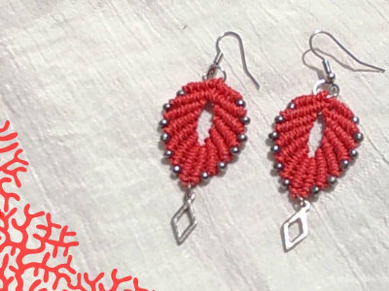 Pode incluir: Um par de brincos vermelhos e prateados com um pingente em forma de diamante. Os brincos s&atilde;o feitos com um cord&atilde;o de n&oacute; e contas prateadas.