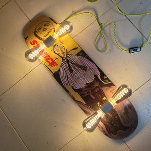Op de afbeelding: Een skateboard hergebruikt als lamp. Het gele skateboard heeft een afbeelding van een man in een pak en het woord "SAUCE". Er zijn drie Edison-stijl gloeilampen bevestigd. De armatuur heeft een gele kabel en een zwarte aan/uit-schakelaar.