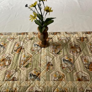 May include: A decorative table runner featuring a repeating pattern of owls, squirrels, and other woodland creatures in hexagonal frames. The runner is beige and green with a quilted design. A small vase with yellow flowers sits on top.