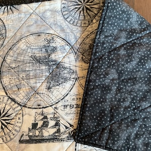 May include: A quilted fabric with a map and compass design in black and white. The fabric features a detailed world map, compass roses, and a sailing ship. The reverse side is a dark gray fabric with white polka dots.