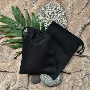 Puede incluir: Dos bolsas de cordón negro hechas de tela, una ligeramente más grande que la otra, están sobre una superficie marrón y peluda. Las bolsas están vacías y tienen un aspecto natural y rústico.