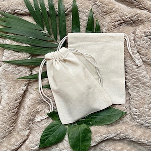 May include: Two natural-colored cotton muslin drawstring bags on a light brown fuzzy surface with green palm leaves.