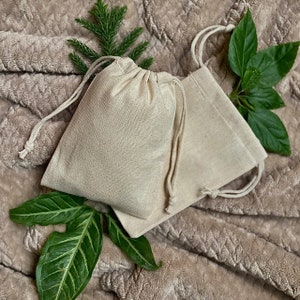 May include: Two natural-colored cotton drawstring pouches with a light brown fuzzy background. The pouches are made of a soft, natural fabric and have a simple, elegant design.