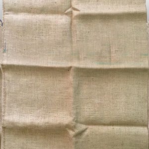 May include: A beige burlap sack with a coarse, woven texture. The sack is rectangular with visible seams and folds, suggesting it is empty. The natural color and material indicate it may be used for storage or rustic decor.
