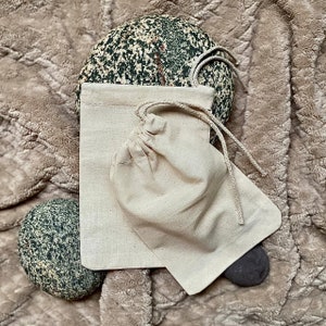 May include: Two natural cotton drawstring pouches with a light brown colour. The pouches are on a brown fuzzy surface with a speckled rock in the background.