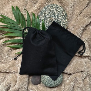 May include: Two black drawstring bags made of fabric, one slightly larger than the other, are lying on a brown fuzzy surface. The bags are empty and have a natural, rustic look.