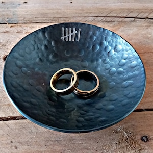 May include: Two gold wedding bands in a black bowl with a hammered texture. The bowl has five vertical lines etched into the surface.