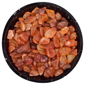 Pode incluir: Vista de cima de uma tigela preta cheia de pedras de cornalina polidas. As pedras variam em tons de laranja e castanho, com uma qualidade translúcida. A borda da tigela proporciona um contraste escuro contra o fundo branco.