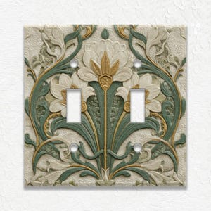 May include: A decorative light switch cover with a floral design. The cover features a raised, textured pattern in cream, gold, and teal. The design includes stylized flowers and leaves, with two switch openings and four screw holes.