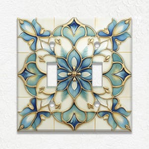 May include: A decorative double light switch cover featuring a floral design in shades of blue, teal, and gold. The design is set against a cream-colored background, with a symmetrical pattern. The cover has two switch openings and a glossy finish.