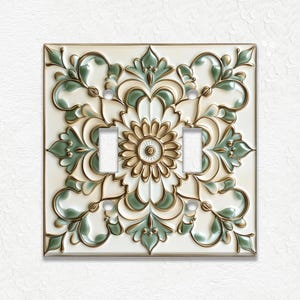 May include: A decorative double light switch cover with a floral design. The cover features a cream-colored background with green and gold accents. The design includes a central flower and surrounding leaves.