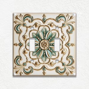 May include: Decorative double light switch cover with a floral design. The cover is white with gold and teal accents. The design is embossed with a raised relief pattern. The switch cover is square and has two switch openings.