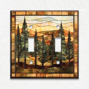 May include: A decorative double light switch cover with a stained glass design. The artwork depicts a forest scene with tall evergreen trees and a mountain range under a warm, sunset-colored sky. The cover has a dark brown border.