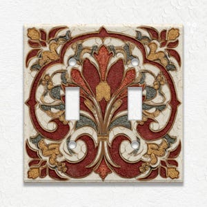 May include: A decorative double light switch cover with an ornate floral design. The cover features a cream-colored background with a detailed pattern in red, gold, and gray. The design includes symmetrical floral elements and scrollwork.