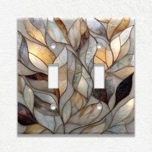 May include: A decorative double light switch cover featuring a stained-glass design of stylized leaves. The leaves are in shades of brown, beige, and gray, with a textured background. The cover has two toggle switch openings and two screw holes.
