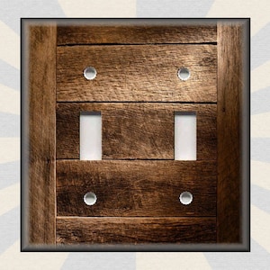 May include: A light switch cover with a rustic wood design. The cover has a dark brown wood grain pattern with a light brown border. The switch plate has two toggle switches and two round decorative accents.
