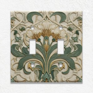 May include: A decorative double light switch cover with an Art Nouveau floral design. The cover features a beige background with raised green, gold, and cream-colored floral and leaf patterns. The switch openings are rectangular.