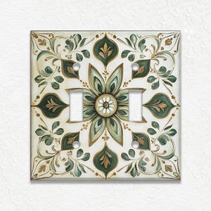 May include: A decorative double light switch cover with a floral design. The cover features a cream background with a green and gold floral pattern. The design includes leaves and a central flower.