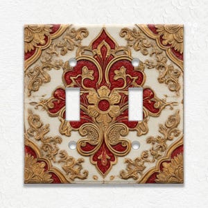 May include: A decorative double light switch cover with an ornate, embossed design. The cover features a cream-colored background with a central red floral pattern and gold accents. The design includes intricate scrollwork and a textured finish.