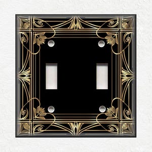 May include: A black light switch cover with a gold Art Deco design. The switch cover has two toggle switches.