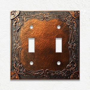May include: A copper-colored double light switch cover with a hammered texture. The cover features an ornate, dark bronze-colored floral border. The switch openings are rectangular, and the screw holes are circular.