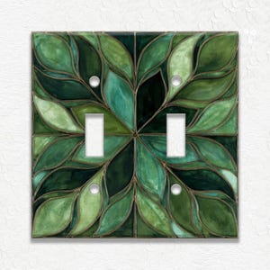 May include: A decorative double light switch cover featuring a leaf design in shades of green. The cover has two toggle switch openings and four small screw holes. The leaf pattern is outlined in a darker green, creating a stained-glass effect.