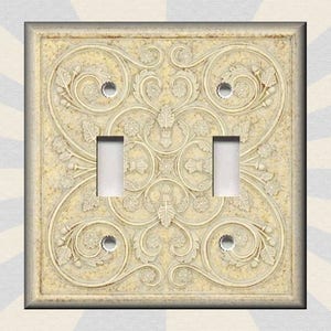 May include: A double light switch cover with a decorative design featuring an intricate pattern of swirls and leaves in a cream color.