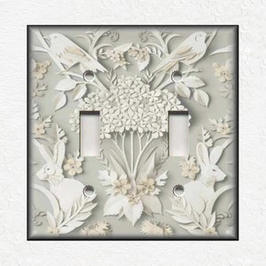 May include: A light gray double light switch cover with a white paper cut design of birds, flowers, and rabbits. The design is intricate and detailed, with a delicate and whimsical feel.