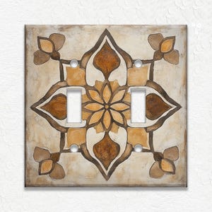May include: A decorative light switch cover with a floral design. The cover features a beige background with a symmetrical pattern in shades of brown and gold. The design includes a central flower and four corner floral accents.