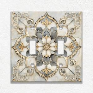 May include: A decorative double light switch cover with a floral design. The cover features a cream, gray, and gold color palette. The design includes a central flower and ornate, symmetrical patterns.