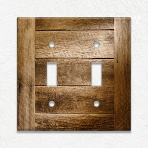 May include: Wooden double light switch cover with a rustic, weathered appearance. The cover features two toggle switch openings and four silver-colored screw holes. The design mimics the look of reclaimed wood planks.