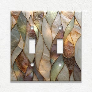 May include: A decorative double light switch cover with a mosaic design. The cover features a pattern of overlapping, leaf-like shapes in shades of gold, beige, green, and brown, outlined with gold. The switch openings are white.