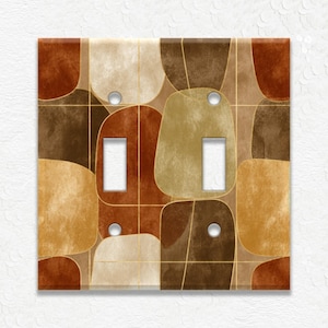 May include: A decorative light switch cover with a mid-century modern design. The cover features a pattern of abstract shapes in shades of brown, tan, and gold, with a grid of thin gold lines. It has two toggle switch openings and two screw holes.