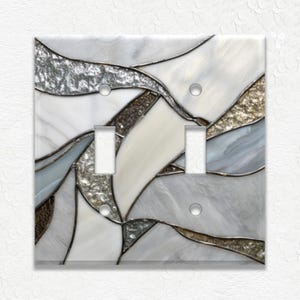 May include: A decorative double light switch cover with a stained glass design. The cover features a mosaic of white, silver, gray, and gold-toned glass pieces, creating an abstract, flowing pattern. The cover has two switch openings and screw holes.