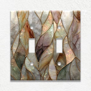 May include: A decorative double light switch cover with a stained glass design. The cover features a mosaic pattern of leaf-like shapes in shades of gray, brown, and iridescent colors, outlined with gold. The switch openings are rectangular.