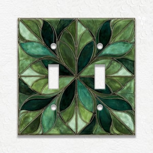 May include: A decorative double light switch cover featuring a stained glass design. The cover showcases a symmetrical pattern of green leaves in various shades, outlined with a dark border. The switch plate has two rectangular openings for the switches and four screw holes.